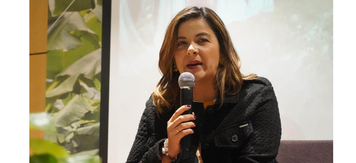 En la imagen se puede ver a la ministra Jhenifer Mojica, con un micrófono en la mano, hablando. De fondo se puede ver un cuadro blanco y al lado unas pantas.