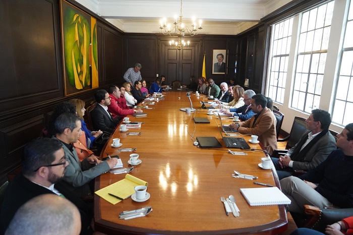La imagen representa a un gran grupo de personas reunidas en torno a una larga mesa de madera, inmersas en una reunión o discusión. En la escena aparecen al menos 14 personas, algunas sentadas y otras de pie alrededor de la mesa. La mesa está llena de diversos objetos, como tazas, tenedores, cuchillos y cucharas, lo que sugiere que los asistentes podrían estar disfrutando de una comida o un refrigerio durante la reunión.