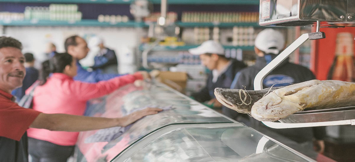 En la imagen se ve a una persona de pie detrás de un mostrador en lo que parece ser un mercado o una tienda de comestibles. La persona lleva una camisa roja e interactúa con un gran pez que cuelga de un anzuelo. El pez es bastante grande y parece ser un tipo de pez de agua dulce, posiblemente una tilapia o una especie similar, dado su tamaño y forma.