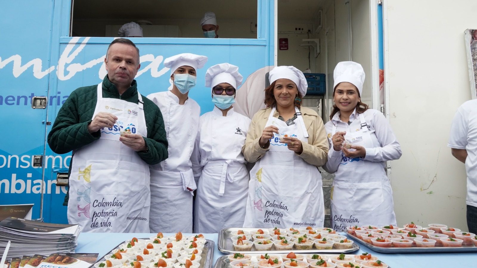 En la imagen aparecen cinco personas de pie delante de una mesa con diversos alimentos.
