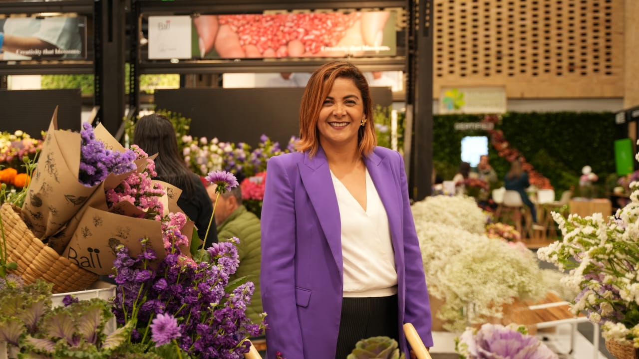 En la imagen aparece la Ministra Jhenifer Mojica de pie en lo que parece ser un mercado de flores o una zona de exposición floral. Lleva una americana morada y un top blanco, y sonríe a la cámara. Detrás de ella hay varias flores y plantas, incluidas flores moradas que probablemente sean lavanda o un tipo de flor similar, así como otras plantas y arreglos florales.