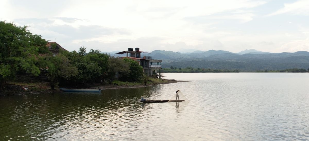 Imagen serena de una masa de agua, probablemente un lago o un río tranquilo, con una persona de pie en una pequeña barca o balsa. La persona parece estar pescando o tendiendo una red, como sugiere la red que sujeta. Al fondo se ve una casa o un pequeño edificio en la orilla, rodeado de exuberante vegetación, lo que indica un entorno rural o semirural.