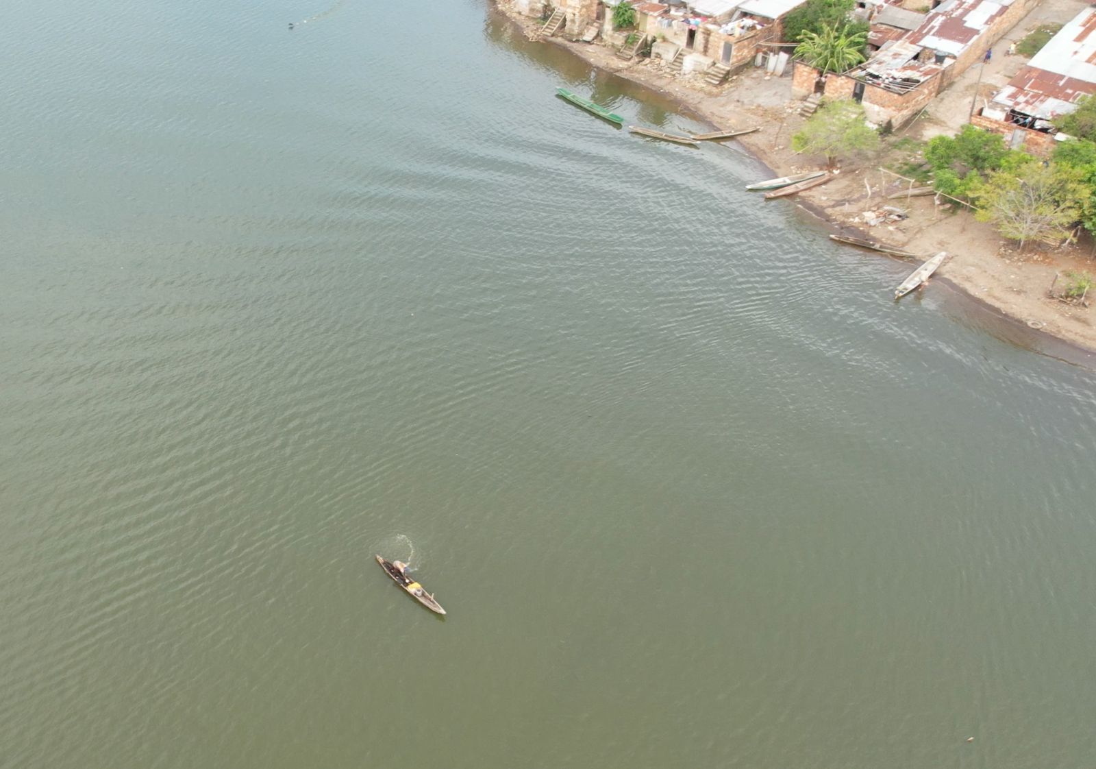 La imagen muestra una masa de agua, posiblemente un lago o un río tranquilo, con una pequeña embarcación en el centro. La embarcación parece ser una canoa o un tipo similar de embarcación, y hay una persona en ella, probablemente remando.
