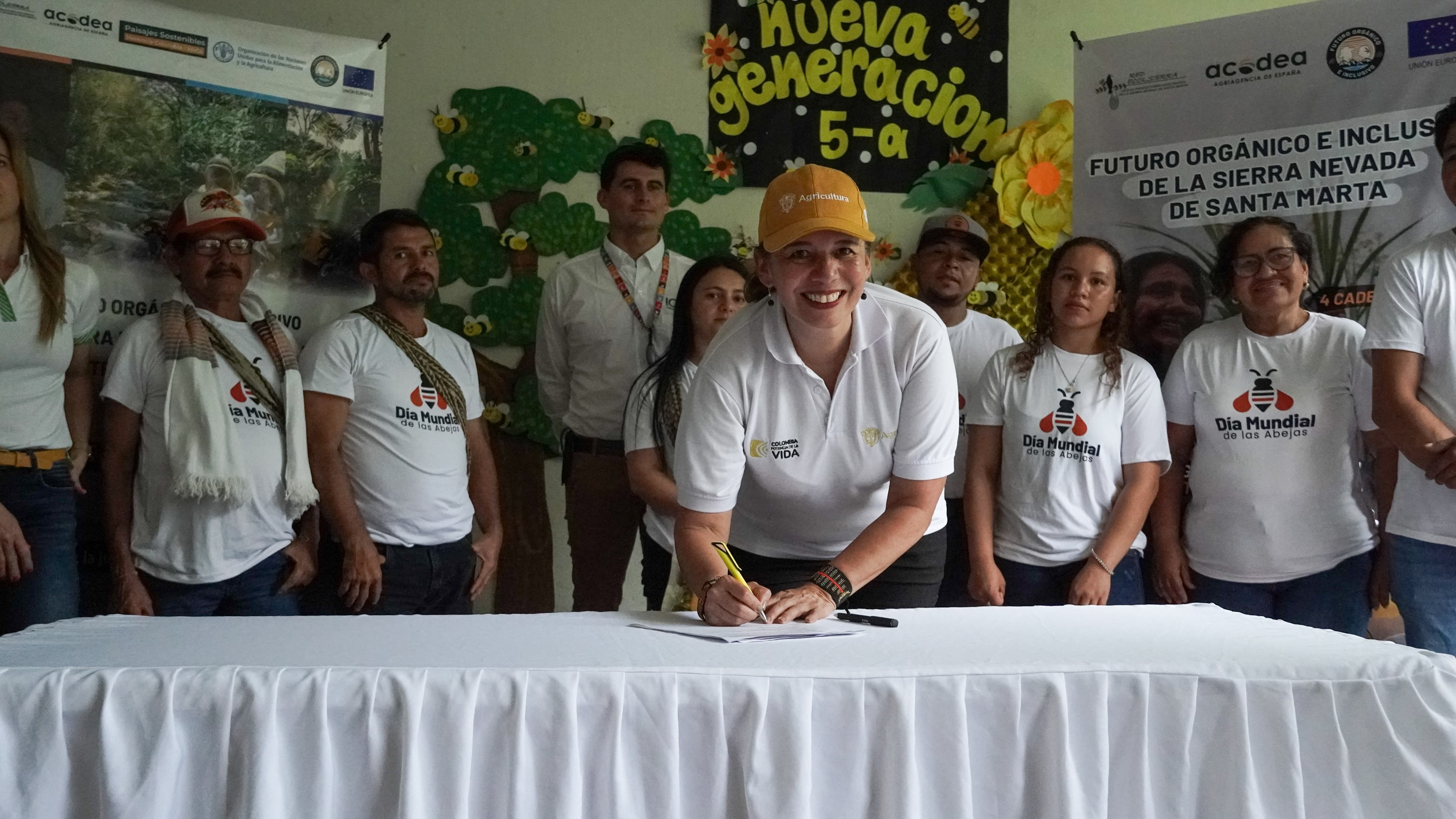 En la imagen, hay un grupo de personas reunidas en torno a una mesa. La figura central parece estar firmando un documento o un libro, práctica habitual en actos formales, ceremonias o la apertura de un nuevo establecimiento. La persona que firma lleva una camisa blanca con un logotipo en el pecho izquierdo y una gorra naranja.