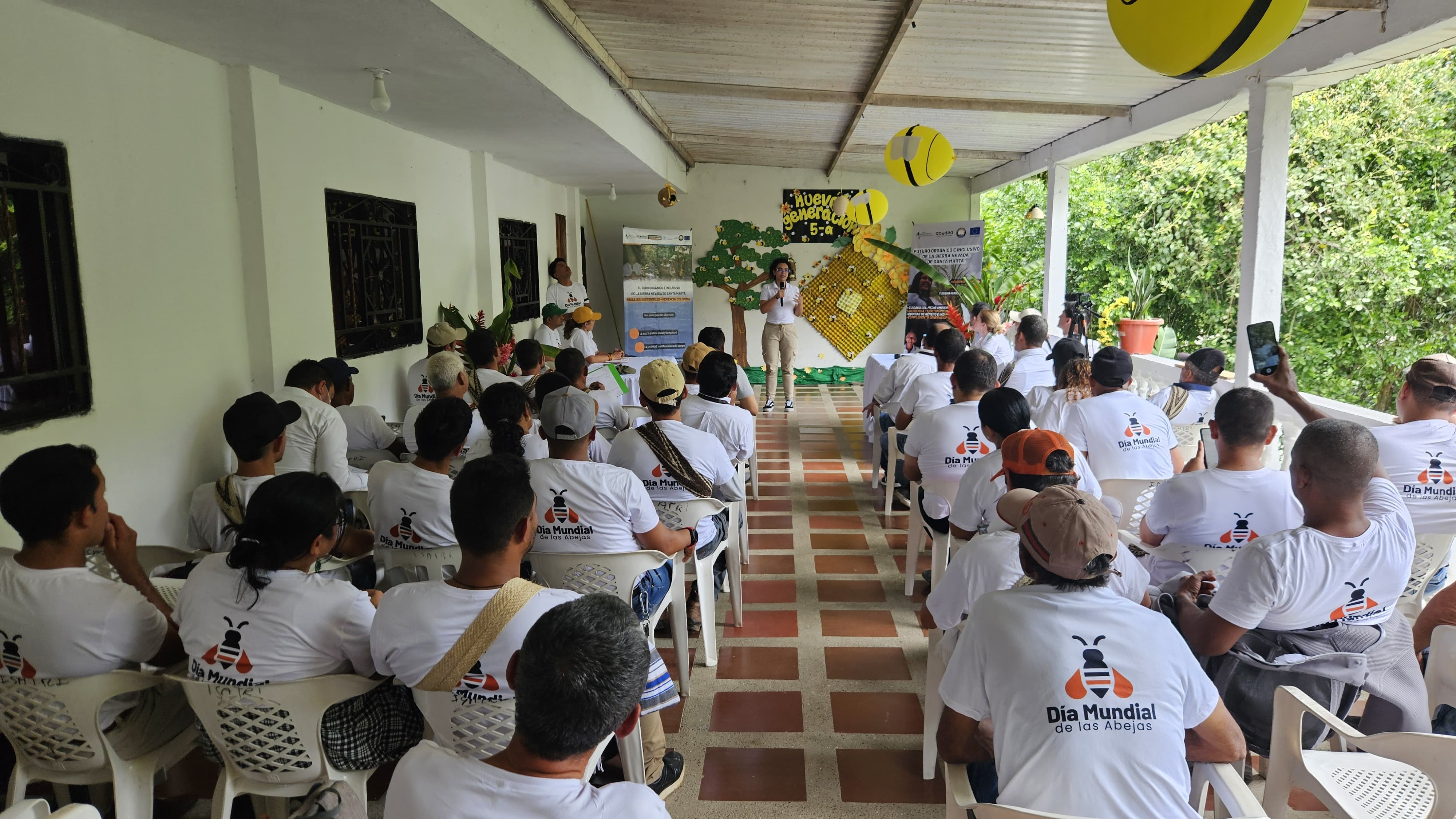 La imagen muestra a un grupo de personas sentadas en filas, mirando hacia el frente, donde parece estar de pie un orador o presentador. El escenario parece un evento al aire libre o semiexterior, posiblemente una reunión comunitaria, un taller o un encuentro. Los asistentes llevan camisetas blancas a juego con un logotipo en la espalda, lo que sugiere que pueden formar parte de una organización o participar en un evento concreto.