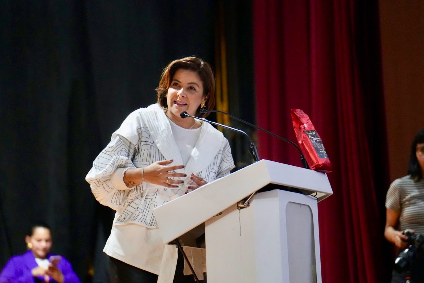 En la imagen aparece la ministra Jhenifer Mojica de pie ante un podio, hablando por un micrófono. Parece estar dando un discurso o una presentación. El escenario parece ser un acto formal o una conferencia, como sugieren el escenario y la presencia de una cortina roja al fondo.