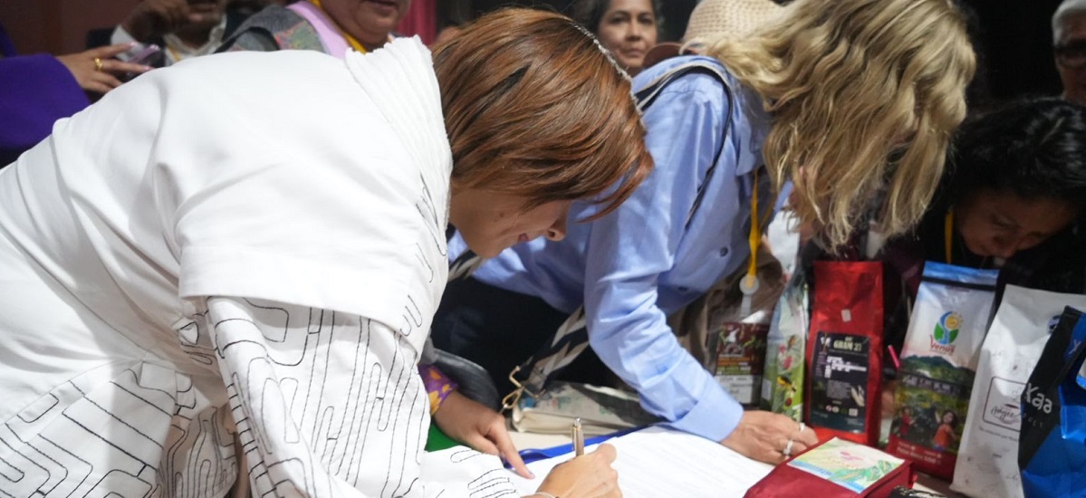 En la imagen se ve a un grupo de personas reunidas en torno a una mesa. Una persona está inclinada sobre la mesa, aparentemente escribiendo o firmando un documento. Esta mujer es la ministra Jhenifer Mojica. 