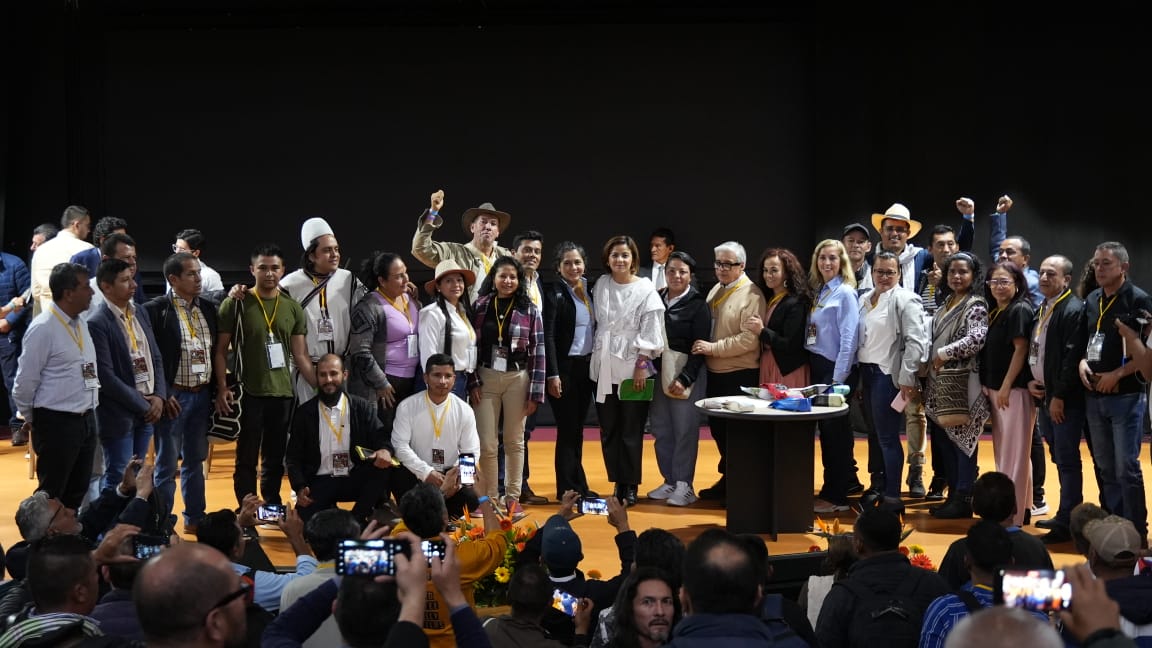 Esta imagen muestra a un grupo de personas reunidas en un escenario, probablemente en un acto o ceremonia. Están en fila, de cara al público, y parecen posar para una foto. El público, en primer plano, está fotografiando al grupo en el escenario, lo que indica que podría tratarse de un acontecimiento importante o de un momento de reconocimiento para las personas en el escenario.