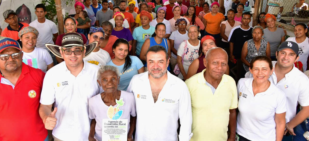 Imagen de un grupo de personas juntas, probablemente en un acto o reunión. Parecen estar posando para una foto, con algunos individuos sonriendo y mirando directamente a la cámara. El grupo es variado, con personas de distintas edades y sexos. Algunas personas llevan sombreros y otra sostiene un cartel o un trozo de papel, lo que podría indicar el propósito de la reunión o la organización a la que pertenecen.