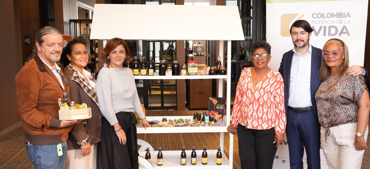 En la imagen aparecen cinco personas de pie delante de un expositor que parece mostrar diversos productos, posiblemente alimentos o bebidas, dadas las botellas y envases visibles. El escenario parece un evento o una exposición en interiores, como indica el telón de fondo con el texto "COLOMBIA POTENCIA DE LA VIDA".