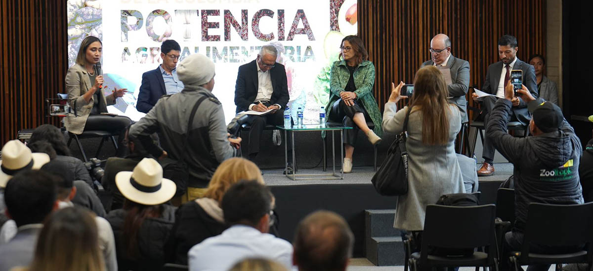En la imagen se puede ver un grupo de seis personas, con lo que parece están presentando un proyecto, algunos de los asistentes a la presentación están tomando apuntes y otros están tomando fotos.