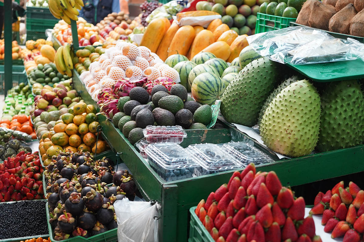Esta imagen muestra un vibrante despliegue de frutas frescas en un mercado. Hay una gran variedad de frutas, como plátanos, naranjas, manzanas, aguacates y lo que parece ser fruta del dragón. Las frutas están dispuestas de forma atractiva, con algunas agrupadas y otras expuestas individualmente. Los colores de las frutas son brillantes y atractivos, lo que sugiere que están frescas y maduras.