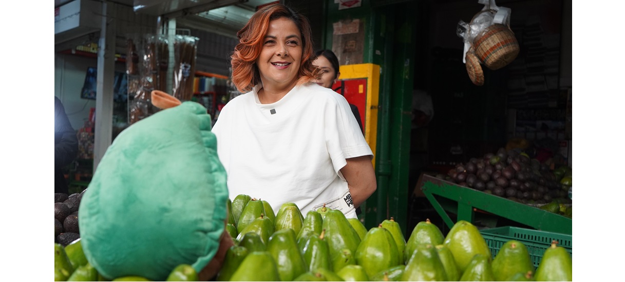 En la imagen aparece la Ministra de pie detrás de un expositor de frutas verdes, que parecen ser aguacates. Sonríe y parece estar en un mercado o tienda, posiblemente vendiendo los productos.