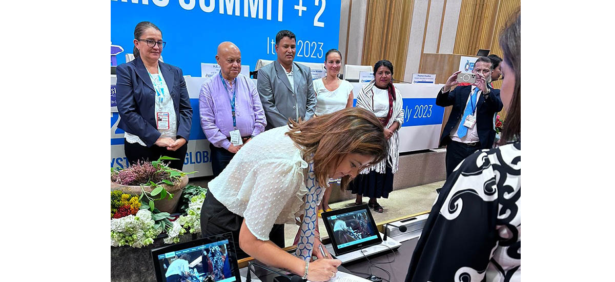 La imagen muestra a una mujer firmando un libro en una mesa, rodeada de varias personas. La mujer lleva una camisa blanca y parece ser el centro de atención. Las personas que la rodean conversan o la observan mientras firma el libro.