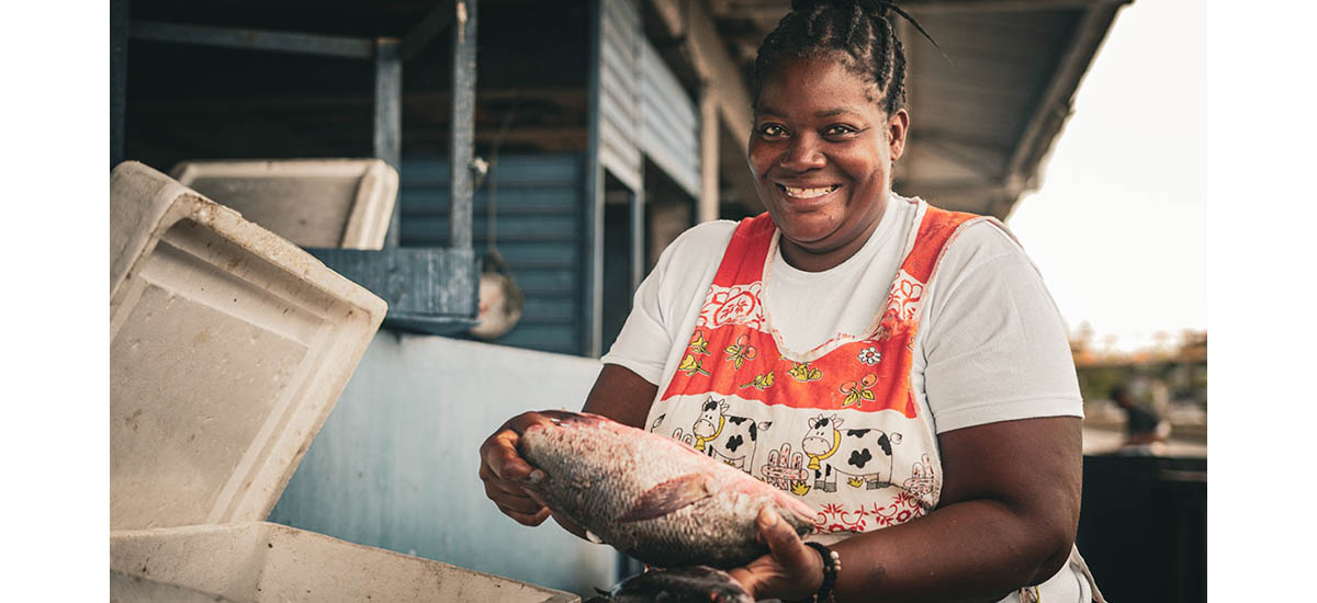 En la imagen se ve a una persona sosteniendo un gran pez. La persona parece sonreír y parece estar en un entorno que podría ser un mercado o un lugar donde se vende o se prepara pescado. El pescado es bastante grande y la persona lo sostiene para mostrarlo.