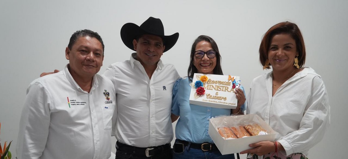 En la imagen aparecen cuatro personas posando para una foto. La persona del centro lleva un sombrero de vaquero y sostiene una caja con lo que parece ser un pastel o un artículo de repostería similar. La caja tiene un diseño colorido e incluye la palabra "Bienvenido". La persona de la derecha es la ministra Jhenifer Mojica quien lleva una blusa blanca y sonríe afectuosamente, mientras que la de la izquierda también sonríe y lleva una camisa blanca con un logotipo en la parte izquierda del pecho. La persona del medio lleva una camisa azul y también sonríe.