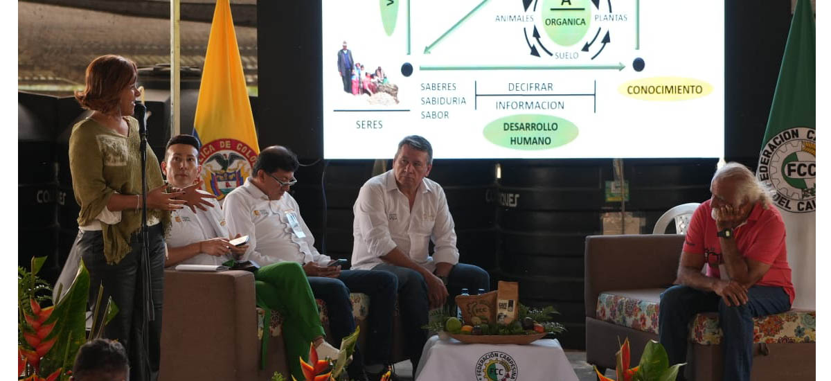 La imagen muestra a un grupo de personas reunidas en una sala, participando en un debate o una presentación. Hay cinco personas en la escena, una de pie cerca del lado izquierdo de la sala, otra en el centro y otras tres a la derecha. Todos están sentados en sillas, con una silla situada en el centro de la sala y otra silla hacia el lado derecho.