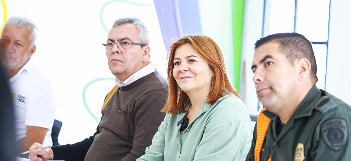 En la imagen aparecen cuatro personas sentadas en lo que parece ser una reunión o conferencia. La persona en primer plano sonríe y mira hacia la cámara, mientras que las otras tres miran en la misma dirección, posiblemente a un orador o a una presentación.