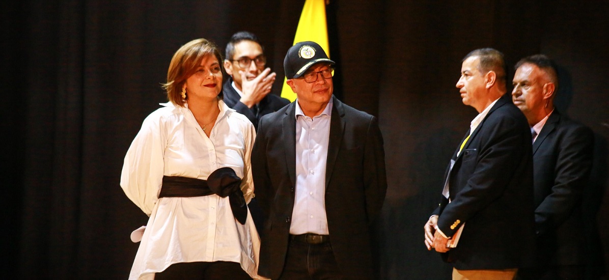 En la imagen se puede ver al presidente Gustavo Petro y a la ministra Jhenifer Mojica, junto a otras personas en un evento o asamblea, de fondo se puede apreciar la bandera colombiana. 