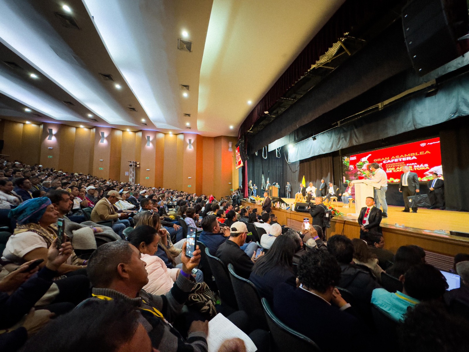 La imagen muestra un acto en un gran salón o auditorio. En la parte delantera hay un escenario en el que varias personas están de pie, posiblemente dando una presentación o actuación.