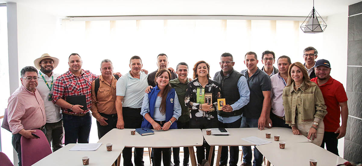 Imagen de un grupo de personas reunidas, probablemente para una reunión o un acto social. Están de pie alrededor de una mesa con algunos papeles y lo que parece ser un libro o una carpeta.
