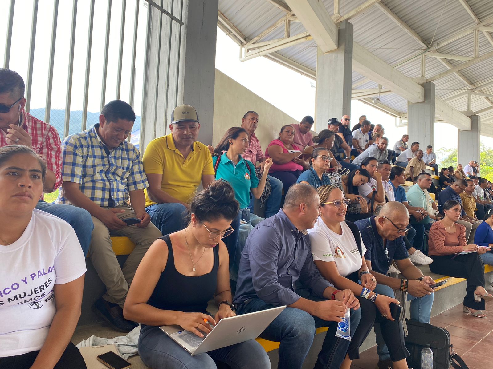 La imagen muestra a un grupo de personas sentadas en lo que parece ser un escenario al aire libre, posiblemente un estadio o un gran espacio abierto. Están viendo algo, probablemente un acontecimiento deportivo o una reunión pública, como sugiere su atención concentrada y la presencia de una gran pantalla o escenario al fondo.