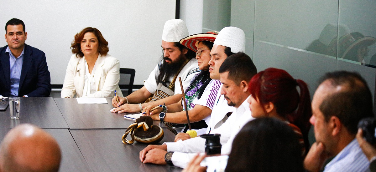 Esta imagen muestra a un grupo de personas sentadas alrededor de una mesa, participando en lo que parece ser una reunión o una discusión. Los individuos están vestidos con una mezcla de atuendos formales y tradicionales, lo que sugiere una mezcla de elementos profesionales y culturales.