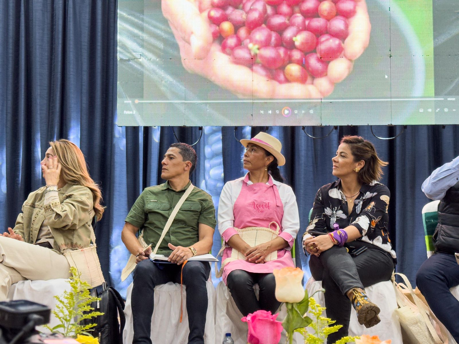 En la imagen aparecen cuatro personas sentadas en un escenario, probablemente en un acto o presentación. La persona del extremo izquierdo parece ser una mujer con el pelo largo, que lleva una chaqueta de color claro y pantalones oscuros.