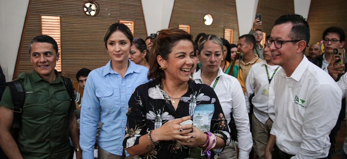 En la imagen se ve a un grupo de personas reunidas, posiblemente en un acto o una reunión social. La mujer del centro sonríe y parece sostener un objeto pequeño, posiblemente un libro o un folleto, y parece ser el centro de atención.