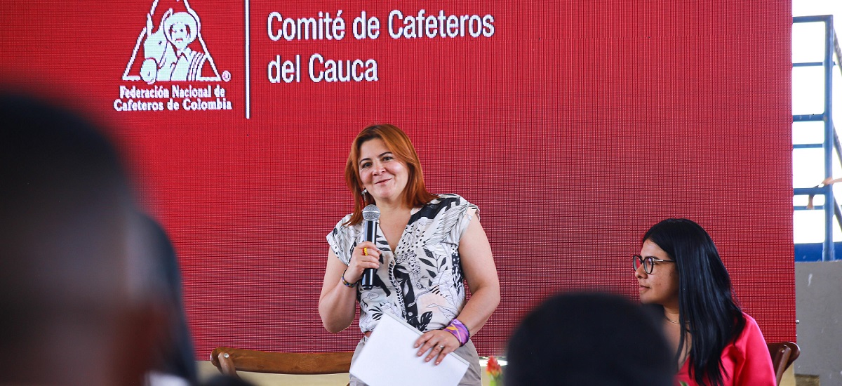 En la imagen, se puede ver a la ministra Jhenifer Mojica de pie delante de un fondo rojo con un logotipo en el que se lee "Comité de Cafeteros del Cauca" y "Federación Nacional de Cafeteros de Colombia". Sostiene un micrófono y parece estar hablando o haciendo una presentación a un público.