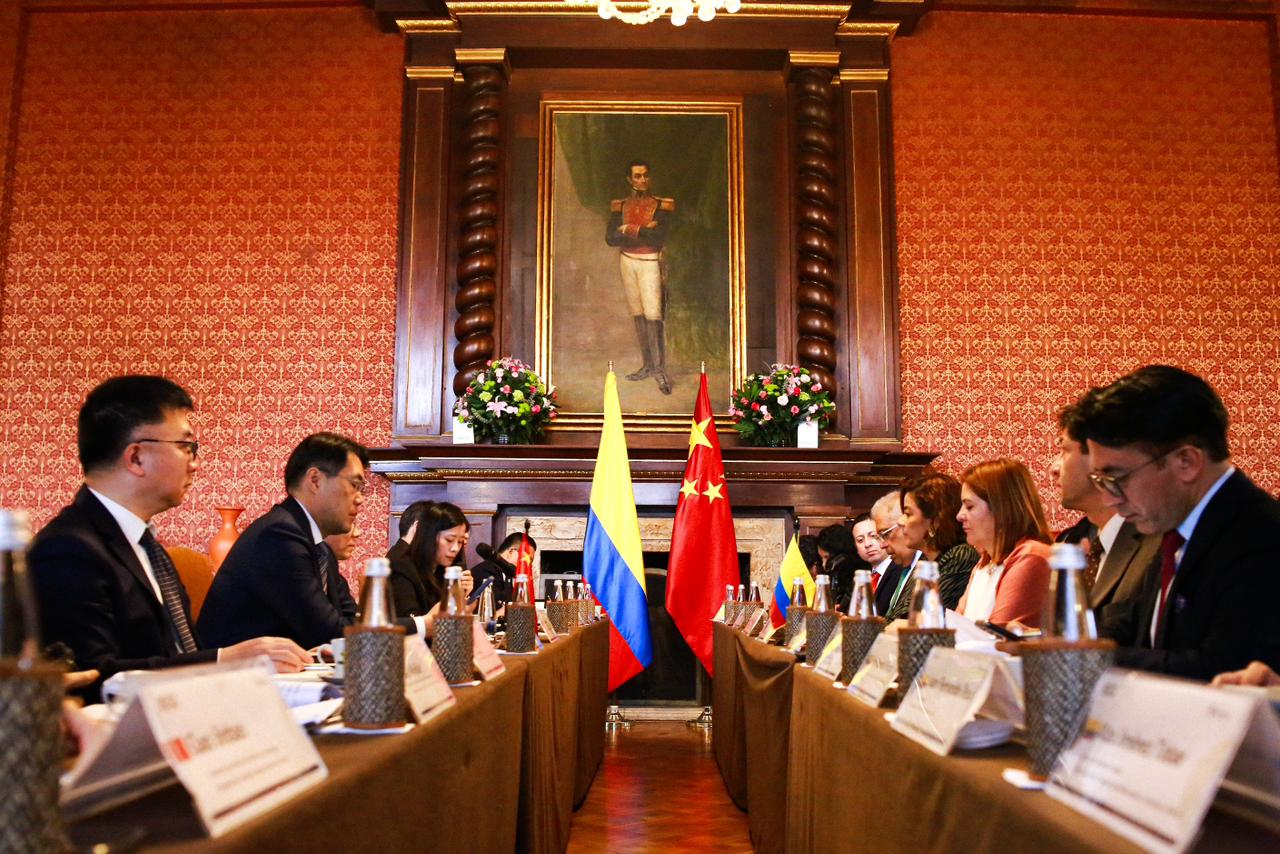 La imagen muestra una reunión formal que tiene lugar en una sala con un interior tradicional y elegante. Hay una mesa larga con varias personas sentadas a ambos lados, frente a frente. Los asistentes parecen estar participando en un debate o una conferencia, como sugiere la presencia de placas con sus nombres delante de ellos y el atuendo formal que llevan.