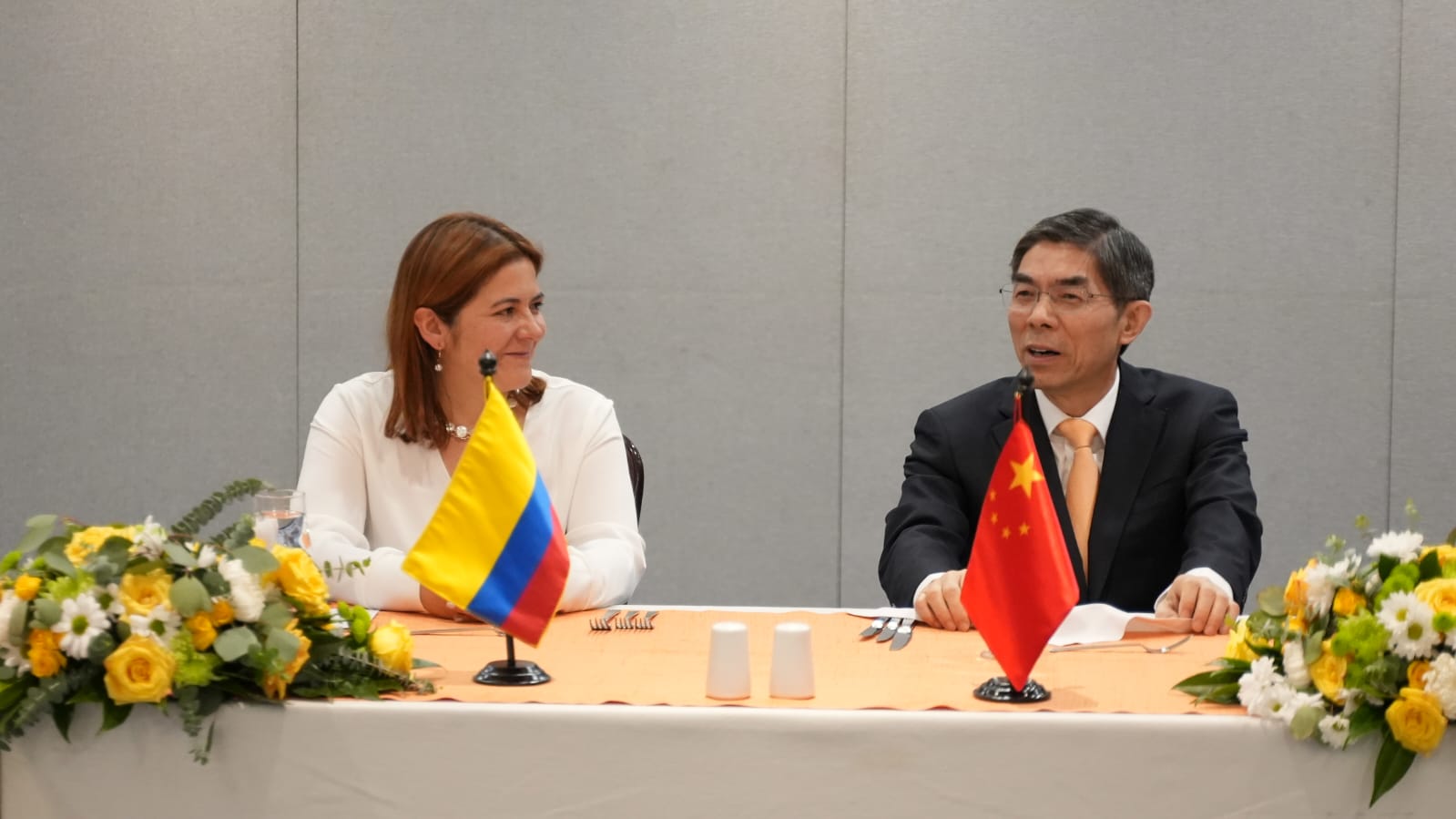 En la imagen aparecen dos personas sentadas a una mesa con un centro de flores. La persona de la izquierda es la ministra Jhenifer Mojica  que lleva un top blanco y sonríe hacia la cámara. La persona de la derecha es un hombre con gafas, traje y corbata, que también mira hacia la cámara.