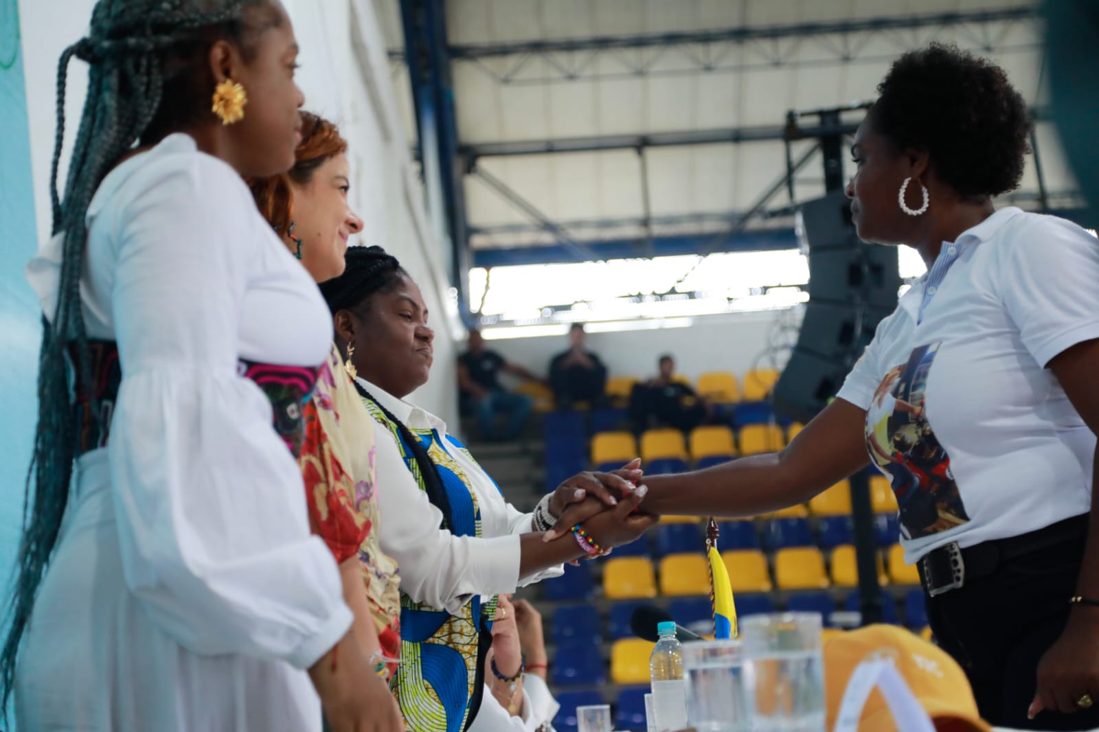 En la imagen aparecen varias personas dándose la mano. La persona de la derecha parece estar estrechando la mano de alguien a quien no se ve bien en el encuadre. El escenario parece un acto o una reunión en un lugar cerrado, posiblemente una ceremonia o una reunión formal, como sugiere la presencia de un escenario y de sillas dispuestas en filas. Las personas están vestidas con una mezcla de trajes formales y semiformales, lo que es habitual en tales ocasiones. El ambiente parece cordial y profesional.