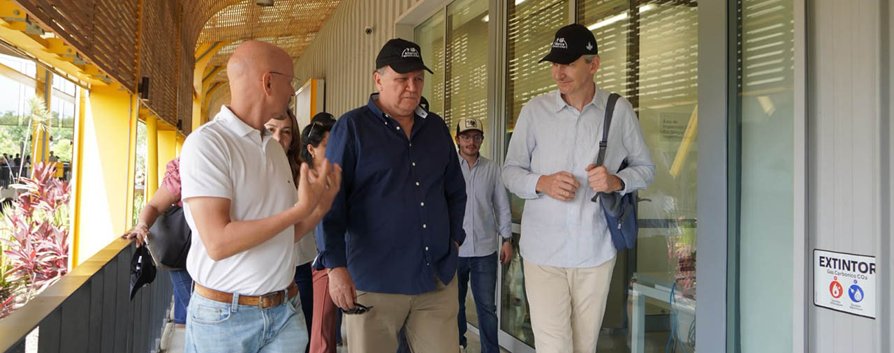 Grupo de personas recorriendo un pasillo cubierto de un edificio con columnas amarillas y ventanas de vidrio, mientras conversan durante una visita o desplazamiento por las instalaciones.