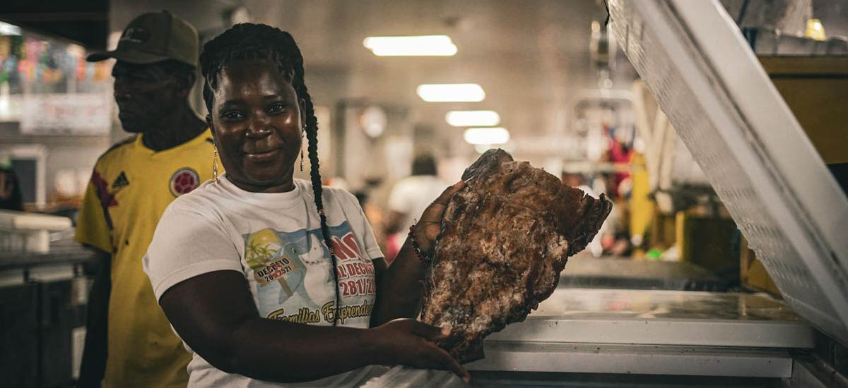 En la imagen aparece una persona sosteniendo un gran trozo de carne, que parece ser un asado o un corte de carne similar. La persona sonríe y parece estar presentando la carne, posiblemente en una cocina o en una zona de preparación de alimentos.