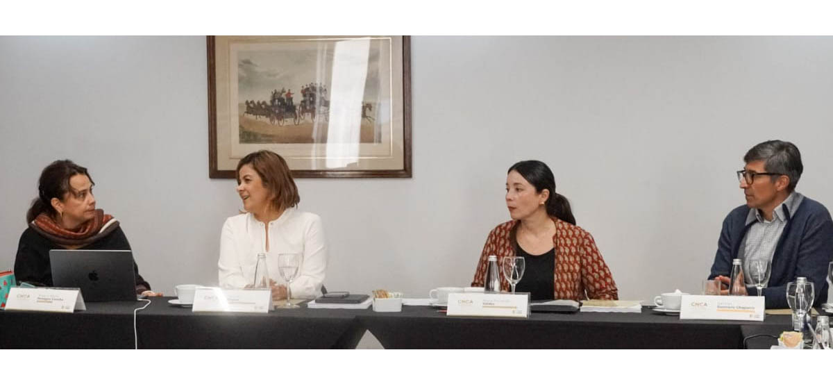 En la imagen, hay cuatro personas sentadas a una mesa, que parece ser un panel o una reunión. Todos visten traje, lo que sugiere un ambiente formal o profesional. También hay portátiles, micrófonos y otros objetos sobre la mesa, lo que refuerza la idea de que se trata de un acto o reunión formal.