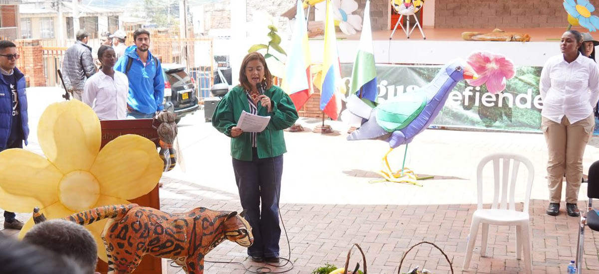 La imagen muestra a una mujer de pie en un podio, dando un discurso a un grupo de personas. Lleva una chaqueta verde y parece ser el centro de atención. El público está formado por varias personas, algunas sentadas en sillas y otras de pie.