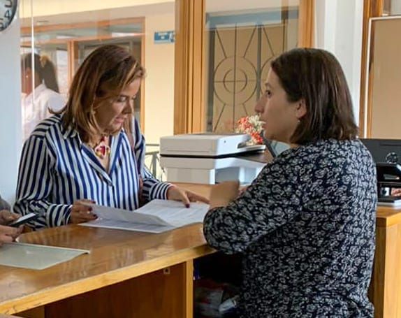 La imagen muestra a tres mujeres de pie frente a un mostrador, enzarzadas en una conversación. Una de las mujeres sostiene un teléfono móvil, posiblemente mostrando algo a las otras dos mujeres. Las mujeres están muy juntas, una a la izquierda del mostrador, otra en el centro y la tercera a la derecha.