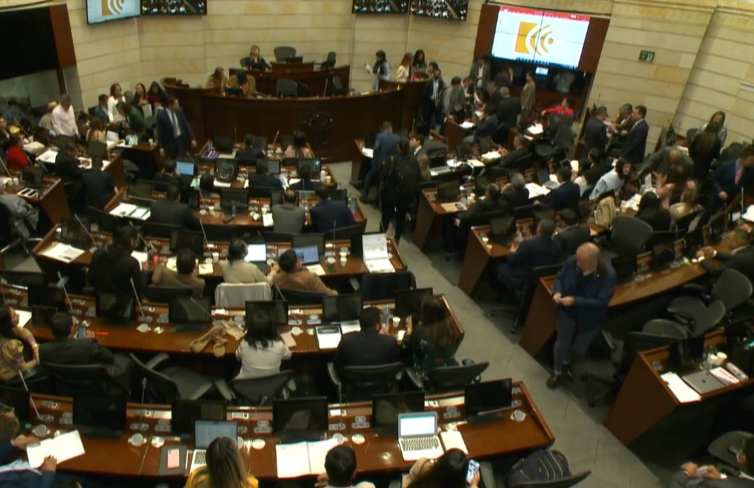 La imagen muestra a un gran grupo de personas reunidas en una sala, posiblemente un parlamento o una sala de conferencias. La sala está llena de personas sentadas en sillas y algunas de pie. Hay varios ordenadores portátiles colocados sobre mesas, lo que indica que los asistentes podrían estar trabajando o discutiendo asuntos importantes.