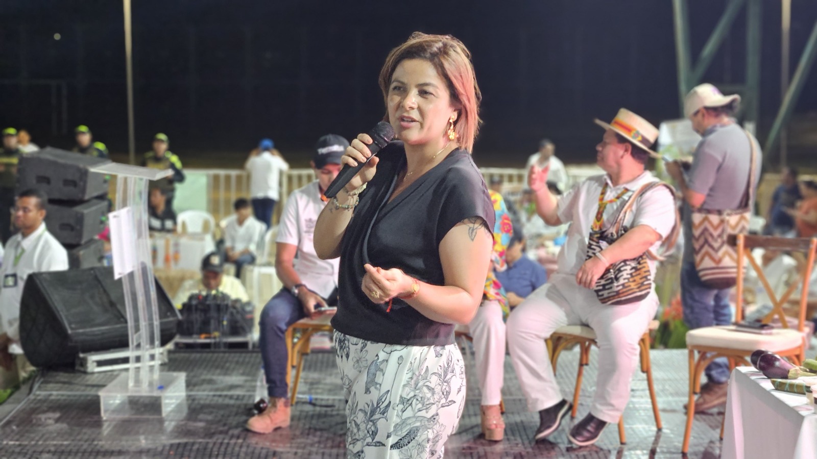 En la imagen aparece la ministra Jhenifer Mojica e pie en un escenario con un micrófono en la mano, Está vestida con un top negro y pantalones estampados, y parece estar hablando con el público. Al fondo hay otras personas, algunas de ellas sentadas en sillas, lo que indica que podría tratarse de un acto o reunión pública.