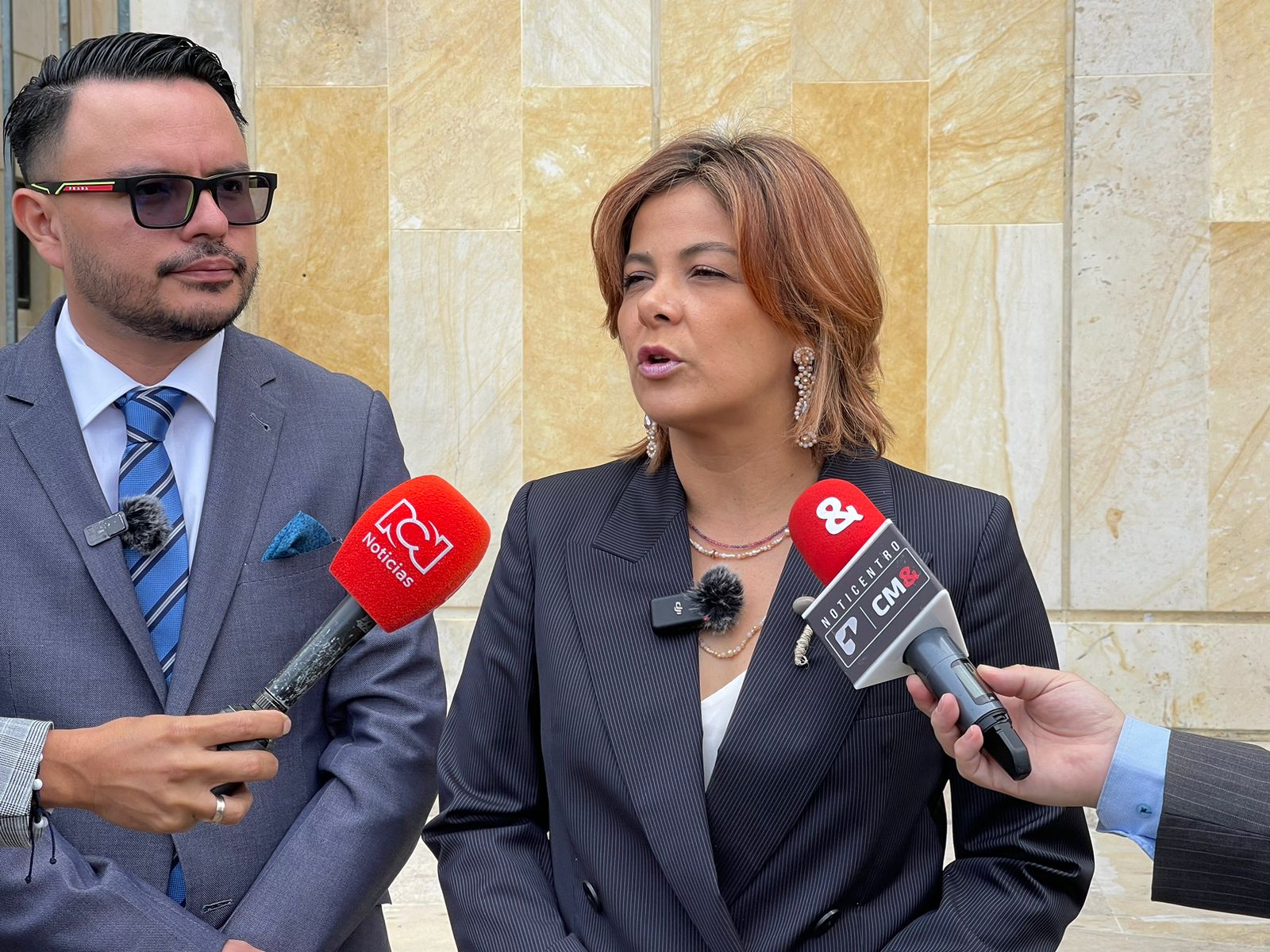 En la imagen hay dos personas que parecen mantener una entrevista con los medios de comunicación. La persona de la derecha habla por un micrófono, lo que sugiere que es la entrevistada. Lleva un traje oscuro y un micrófono enganchado a la chaqueta, lo que indica que es el sujeto de la entrevista. La persona de la izquierda está de pie a su lado, lleva traje y gafas de sol, y parece estar escuchando la entrevista.