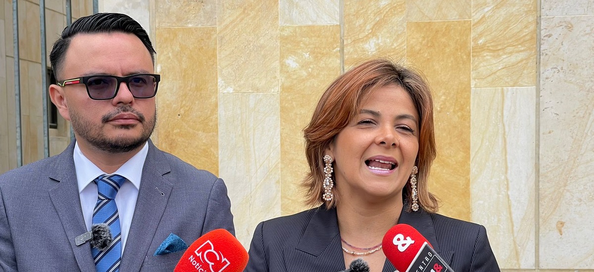 En la imagen hay dos personas que parecen estar participando en un acto con los medios de comunicación o en una rueda de prensa. La persona de la izquierda es un hombre que lleva gafas de sol, traje y corbata, y tiene un micrófono enganchado a la chaqueta, lo que sugiere que puede estar hablando o a punto de hablar. La persona de la derecha es una mujer con el pelo corto, que lleva traje y pendientes, y también tiene un micrófono delante, lo que indica que probablemente sea una oradora o entrevistada.