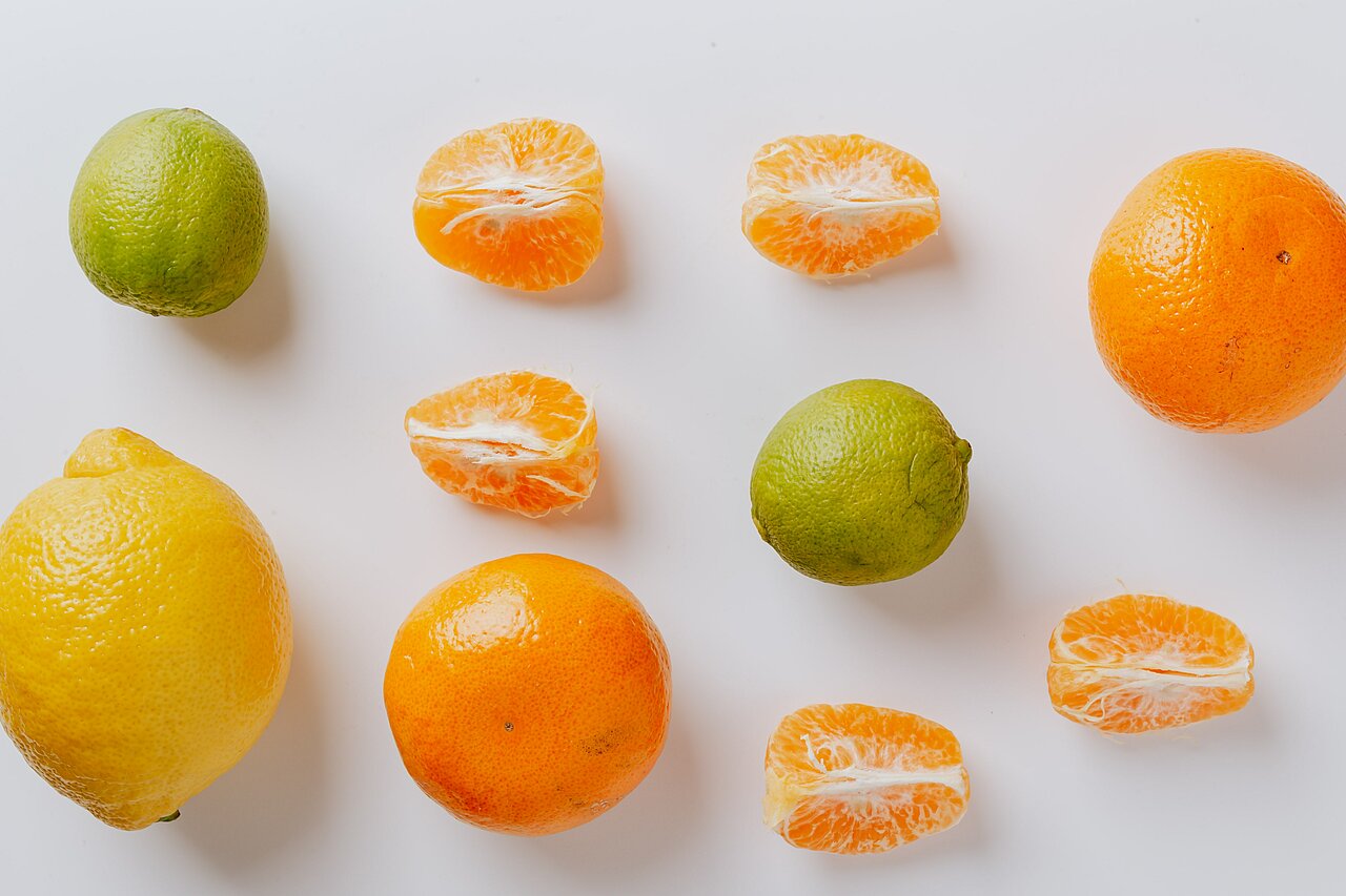 La imagen muestra un fondo blanco con diversos cítricos dispuestos de forma visualmente atractiva. Sobre la mesa hay varias naranjas y limas, algunas de ellas cortadas en rodajas. Las naranjas son de distintos tamaños y están repartidas por toda la mesa, mientras que las limas están colocadas más cerca del lado izquierdo de la mesa. La disposición crea una colorida y acogedora muestra de cítricos frescos.