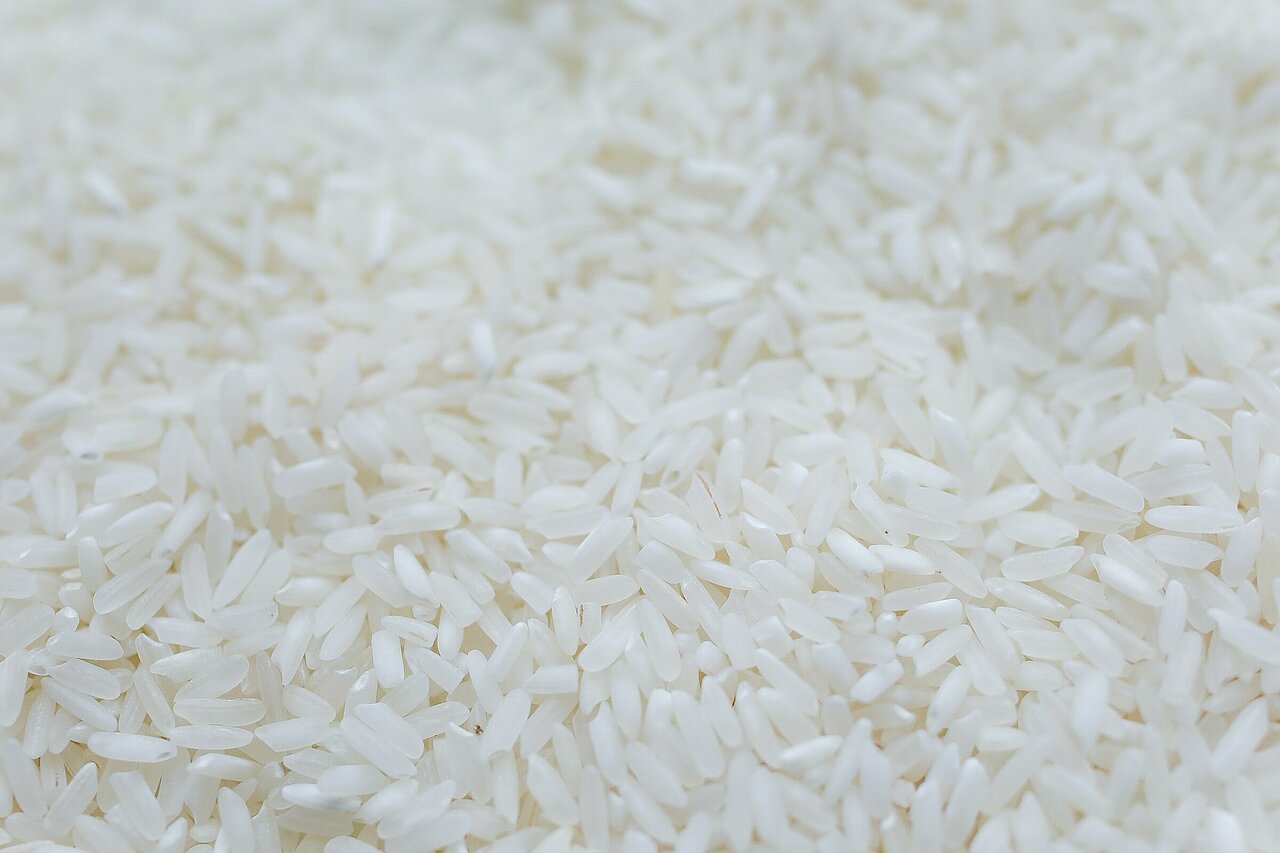 La imagen muestra un gran montón de arroz blanco, que parece estar crudo. El arroz está extendido en una capa plana y uniforme que cubre todo el encuadre de la imagen. El primer plano del arroz destaca su textura y la uniformidad de los granos. La escena sugiere que el arroz está listo para ser cocinado y preparado para una comida.
