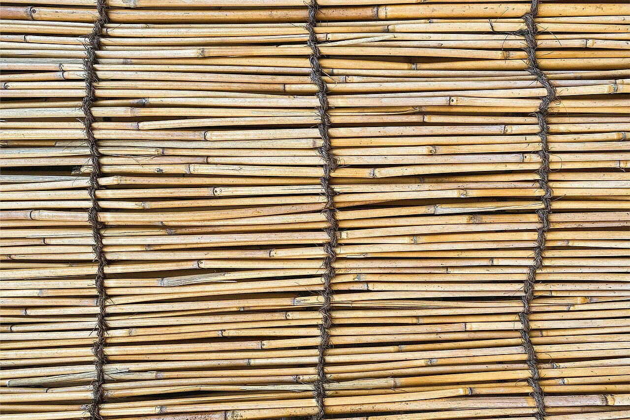 La imagen muestra una pared hecha de guadua, creando un diseño único y natural. Las varas de guadua están dispuestas siguiendo un patrón, con algunas superpuestas y otras colocadas horizontal o verticalmente. La pared está formada por varias capas de guadua, lo que le da un aspecto texturado y visualmente interesante. Los palos varían de tamaño y orientación, lo que añade profundidad y complejidad al diseño.