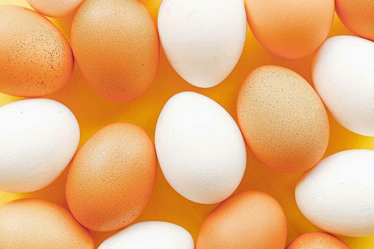 La imagen muestra una gran pila de huevos, con una mezcla de blancos y marrones. Los huevos están dispuestos de una manera visualmente atractiva, con algunos colocados unos encima de otros, mientras que otros están esparcidos. La escena crea una sensación de abundancia y variedad, mostrando los diferentes colores y tamaños de los huevos. La disposición de los huevos sobre la mesa constituye una atractiva muestra, probablemente destinada a la venta o a la decoración.