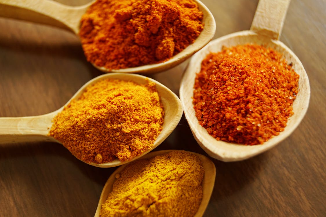 La imagen muestra tres cucharas de madera llenas de diversas especias, colocadas sobre una mesa de madera. Las especias son amarillas, naranjas y rojas, lo que indica una gran variedad de sabores. Las cucharas están dispuestas de forma que se vean las distintas especias, lo que facilita su elección y uso para cocinar o condimentar.