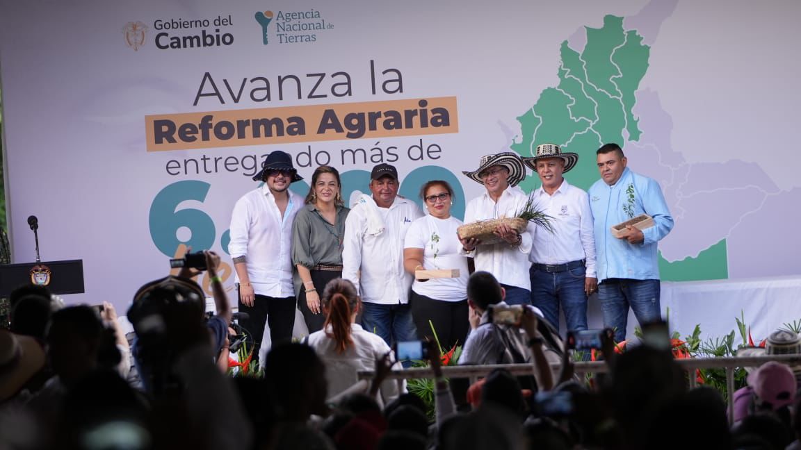 La imagen muestra a un grupo de personas de pie en un escenario, probablemente en un acto o ceremonia. Visten trajes formales y algunos llevan sombreros, lo que sugiere un significado cultural o tradicional de la ocasión. El telón de fondo muestra un mapa y un texto que dice «Avanzamos la Reforma Agraria» y «Gobierno del Cambio».