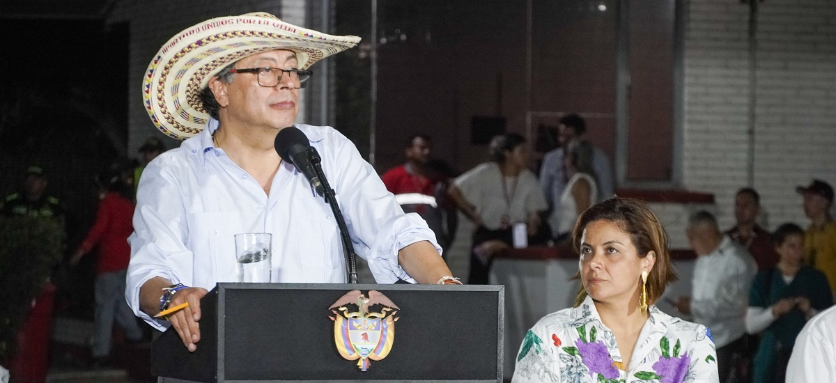 En la imagen aparece el presidente Gustavo Petro, de pie ante un podio, hablando por un micrófono. Lleva sombrero de paja, gafas y camisa clara. Detrás de él, la ministra Jhenifer Mojica escuchando atentamente al presidente.