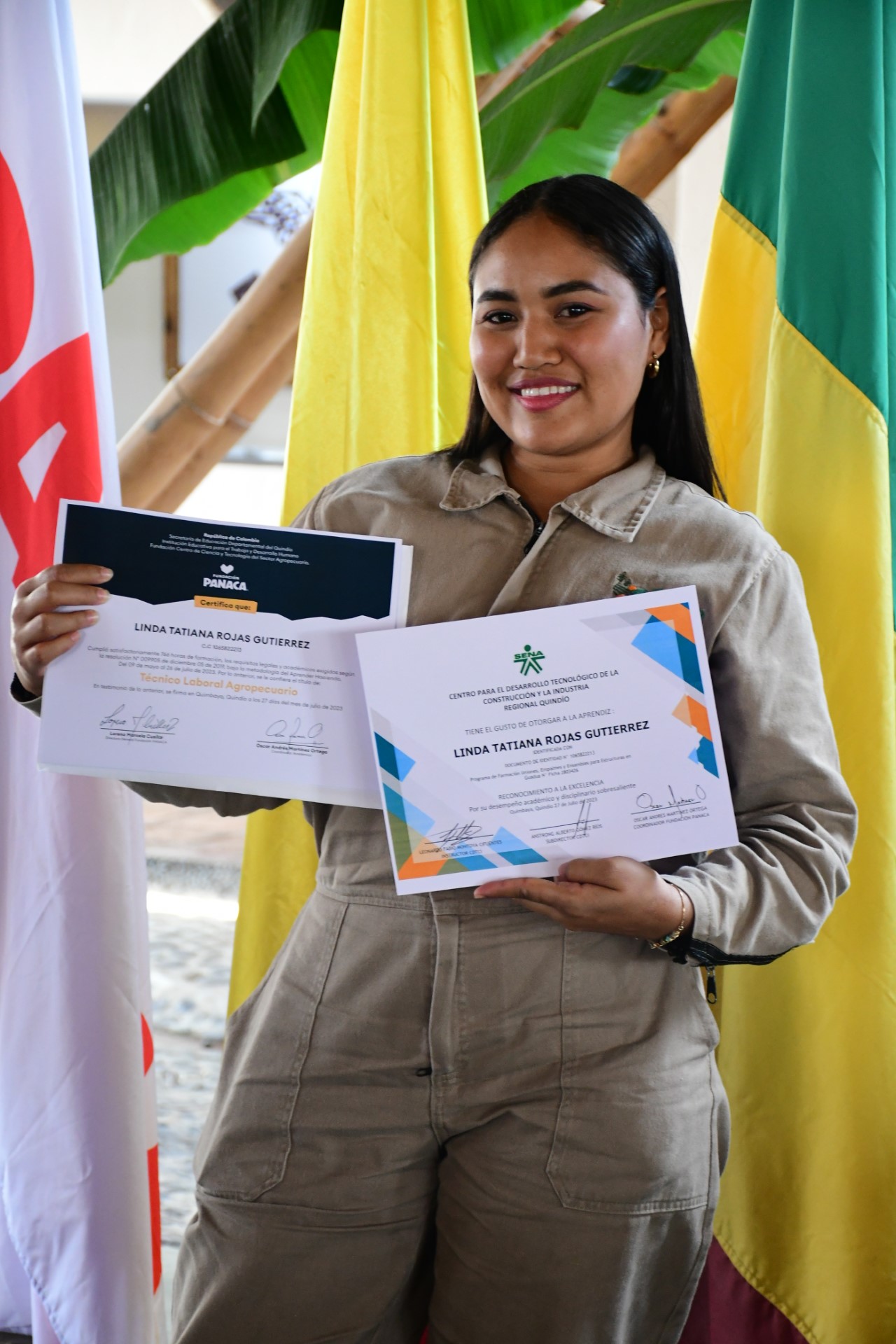 La imagen muestra a una mujer de pie delante de una bandera, con dos certificados en las manos. Parece mostrar con orgullo los documentos, posiblemente indicando sus logros o realizaciones. La mujer lleva uniforme, lo que sugiere que podría ser una profesional o miembro de una organización.